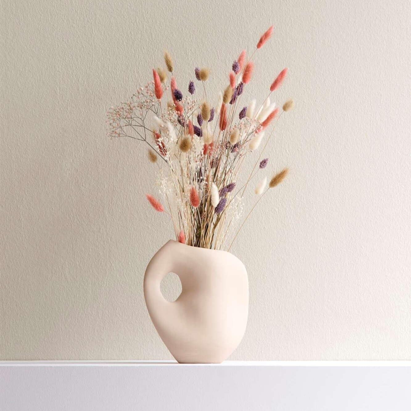 Dried floral arrangement in a modern ceramic vase with a round cutout on a white surface.
