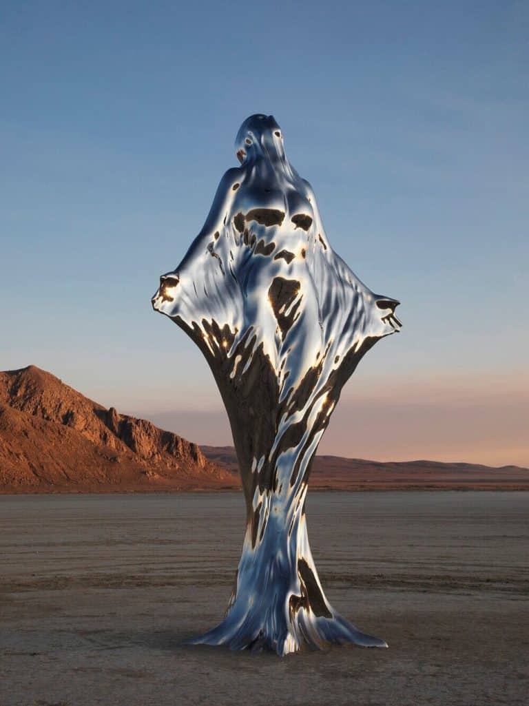 Michael Benisty Creates Stunning And Majestic Sculpture Installations Reflective metal sculpture of a woman in desert landscape at sunset.