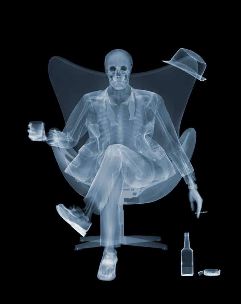 Interview With Nick Veasey X-ray image of a person sitting in a chair with a drink, hat, and cigarette, with a skeletal appearance and relaxed pose.