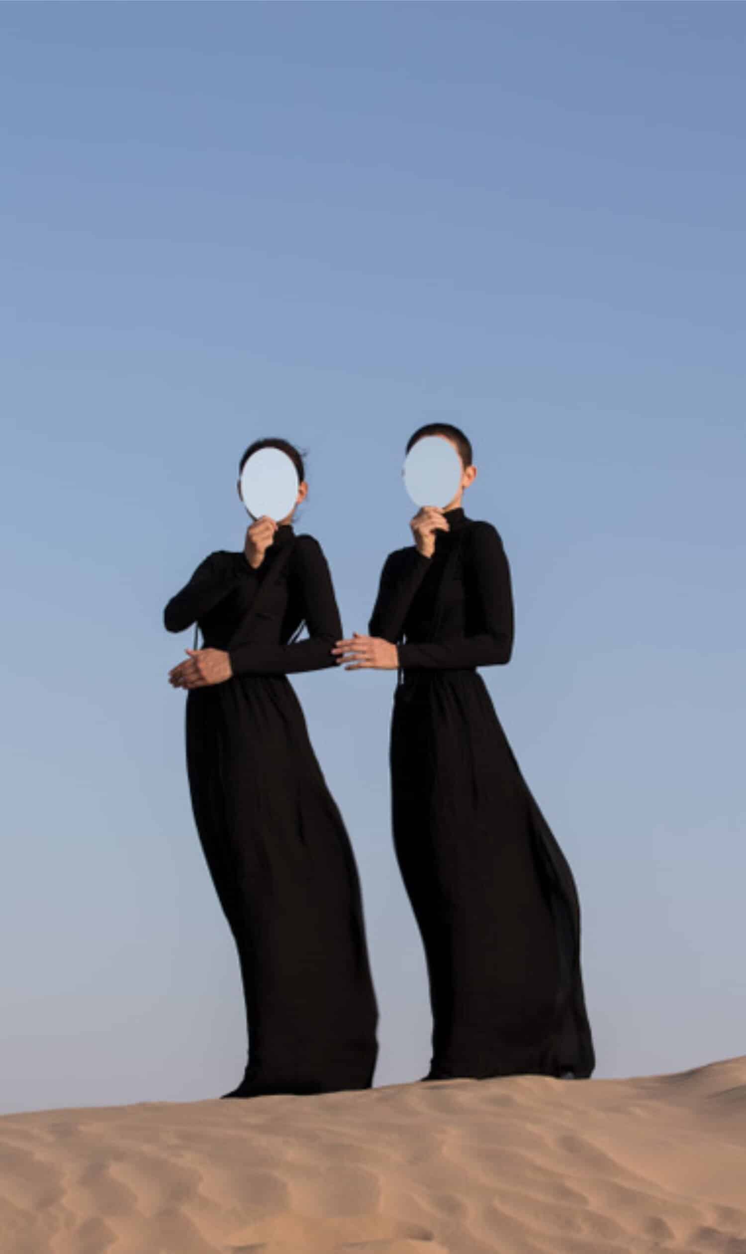 Ethereal women in black dresses holding mirrors over desert landscape against clear blue sky.
