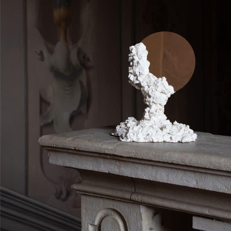 Davide Ronco Creates A Family Of Botticino X Palazzo Monti Furniture Weathered sculpture of a face on a stone pedestal with a circular mirror background.