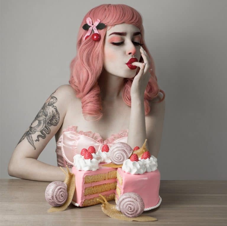 Petite Doll Creates Radiant Allusions Of A Surreal Time And Place Vibrant pink-haired woman with tattoo and vintage style dress celebrating with a colorful cake.