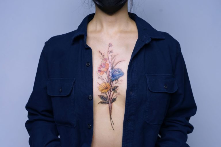 Geunhee's Tattoo Artistry: A Blossoming Tribute to Family Love and Resilience Colorful floral tattoo on woman's chest with dark shirt and face mask.