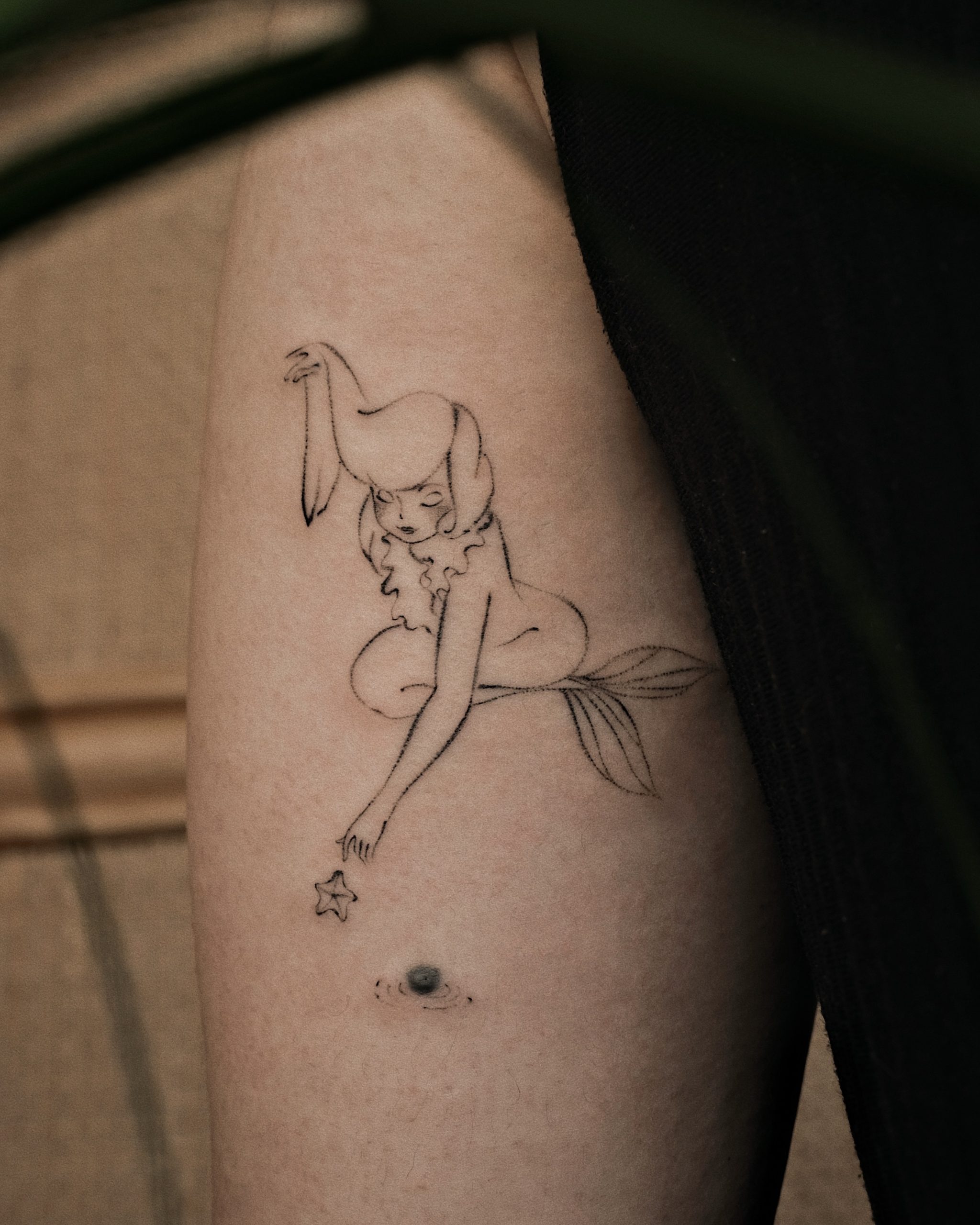 Delicate line art tattoo of a mermaid with a pony on her head and leaf tail on arm, black ink on light skin.