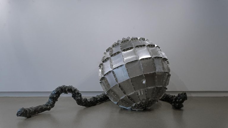 Xinyu Zhang is Crafting Contemporary Dialogue Through Sculpture Chromed geometric sculpture resembling an atom or virus structure placed on a neutral surface.