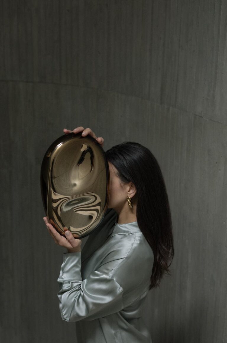Milena Kling introduces new ethereal mirror sculptures Reflective bronze art sculpture held by woman with dark hair and gold earrings.