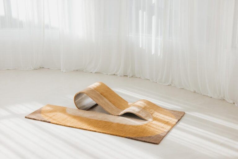 Budde and Nando Studio transform the rug into a sculptural chaise lounge Soft beige and gray modern abstract area rug with a metal sculptural element, set in a brightly lit minimalist room.