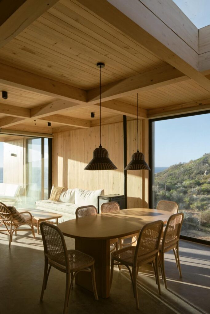 Interior shot highlighting the open-plan living and dining areas with wooden beams, minimalist furniture, and expansive windows facing the ocean