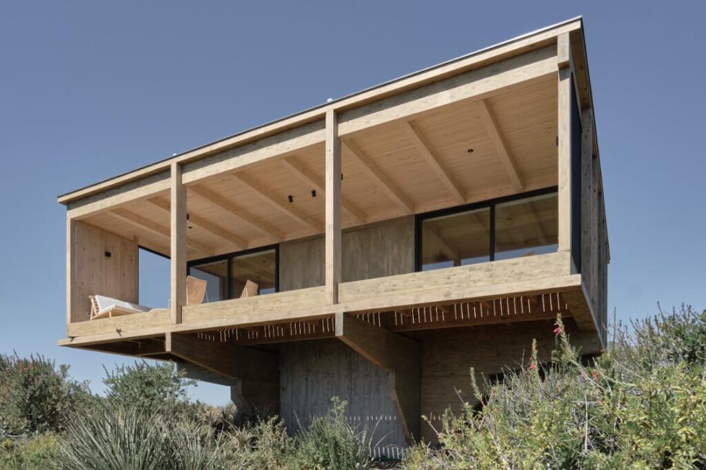 Elevated exterior view of CR House showing its wooden quadrants and central concrete core amid coastal vegetation and rugged terrain