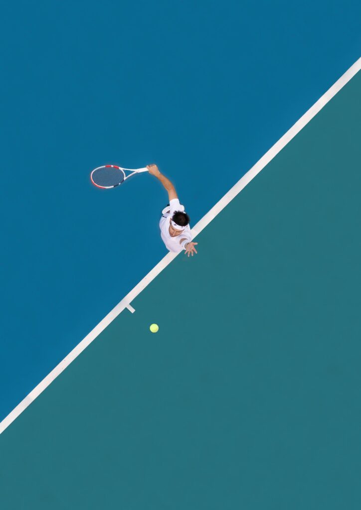 Aerial view of a tennis player mid-serve on a blue hardcourt with sharp shadow contrast