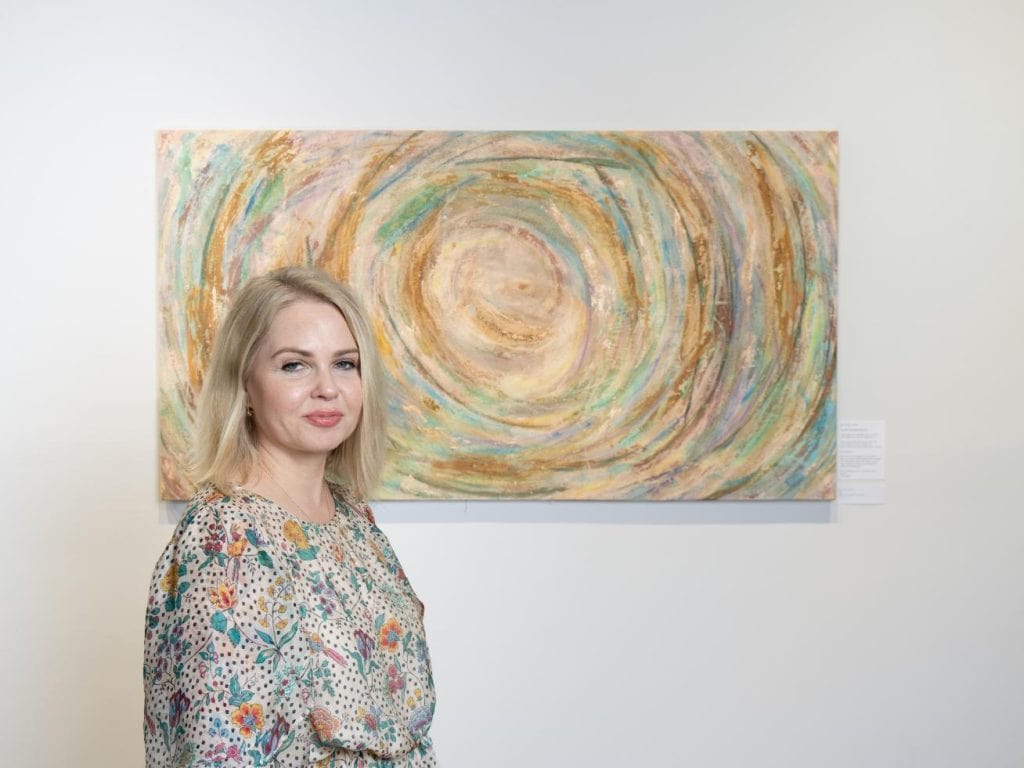 The artist poses beside one of her pieces, her expression calm and reflective, embodying the spiritual nature of her practice.