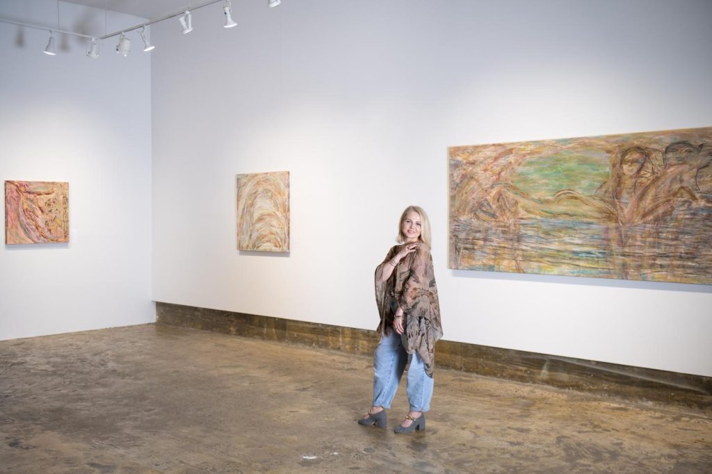 Kasia Muzyka stands in a softly lit gallery, gazing at one of her large abstract works that blends flowing forms and luminous colors.