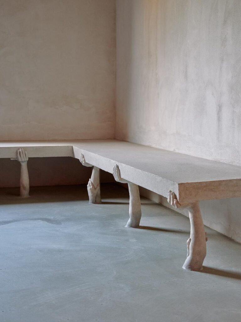 Bench-like structure supported by sculptural legs and hands emerging from the floor