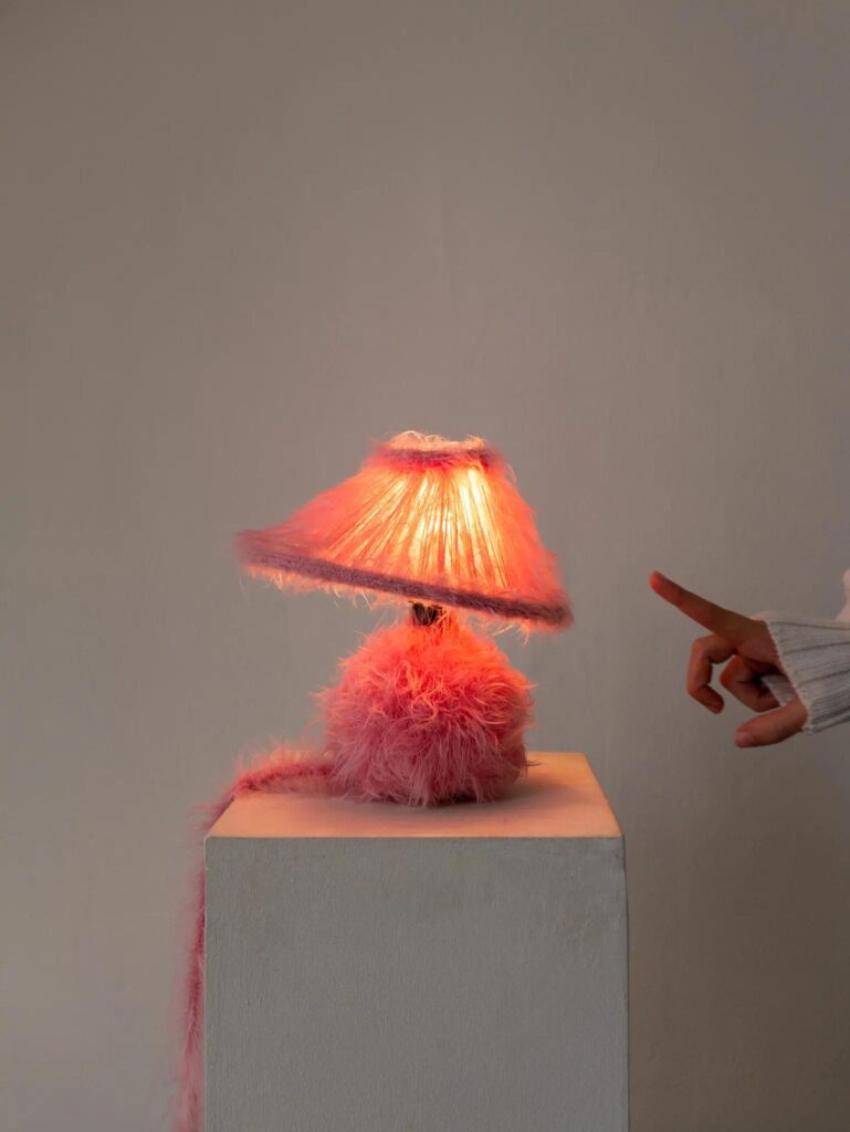 Pink furry table lamp by napalosa_ lit on a white pedestal with hand entering frame