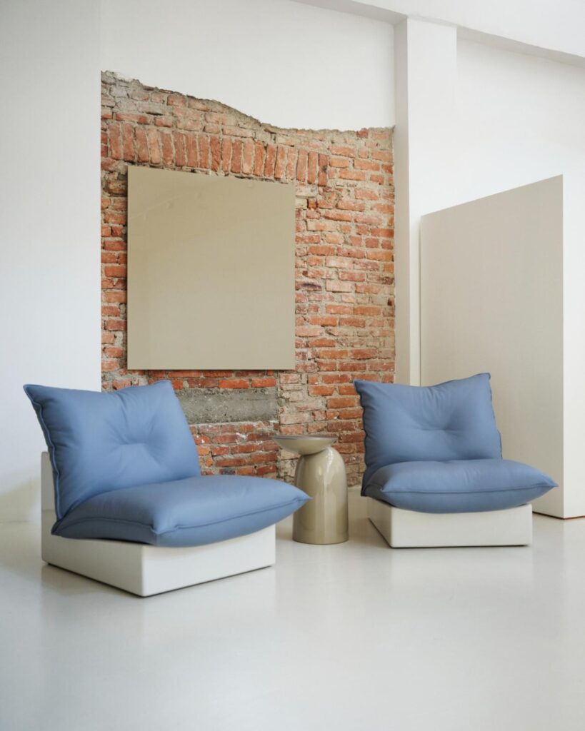 Two low lounge chairs with blue cushioned seats against an exposed brick wall with a small side table